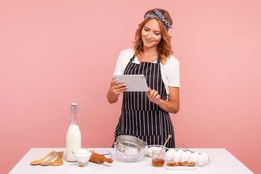 Housewife chef cook confectioner or baker searching new recipe in internet, standing with tablet in hands near table with products for baking. Indoor studio shot isolated on pink background.