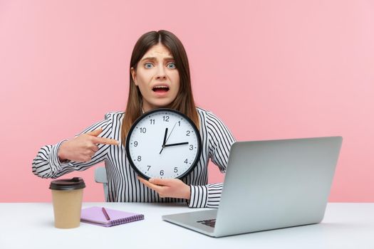 Nervous anxious woman office worker pointing finger at big wall clock in her hands sitting at workplace, working late hours, overtime job. Indoor studio shot isolated on pink background