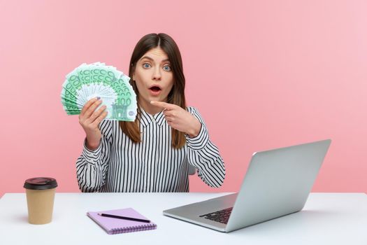 Surprised excited woman office worker pointing finger at hundred euro bills sitting at workplace looking with astonishment, shocked with high salary. Indoor studio shot isolated on pink background