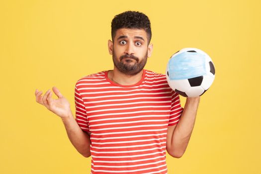 Frustrated young bearded man in red t-shirt holding in hand soccer ball in medical mask, problems with the football championship, corruption. Indoor studio shot isolated on yellow background