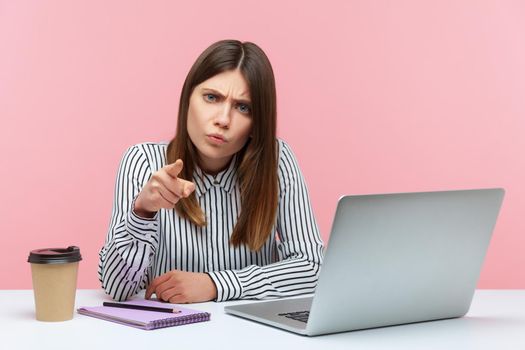 Hey you. Irritated angry businesswoman in striped shirt pointing finger to camera, scolding and making you guilty, sitting at workplace home office. Indoor studio shot isolated on pink background