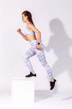 Full length side view persistent athletic woman in white sportswear doing cardio exercises, stepping on cube, developing strength and endurance. Indoor studio shot isolated on gray background