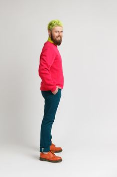 Side view of happy bearded man expresses positive emotions, looks at camera with toothy smile, wearing red sweater and elegant pants. Indoor studio shot isolated on gray background.