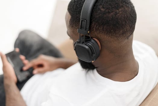 African american young male enjoying music in wireless headphones while sitting on sofa at home. Close up.