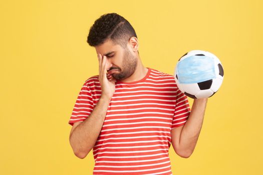 Deeply upset bearded man crying holding ball in protective mask in hand, want to visit championship, closed matches because of coronavirus covid-19. Indoor studio shot isolated on yellow background