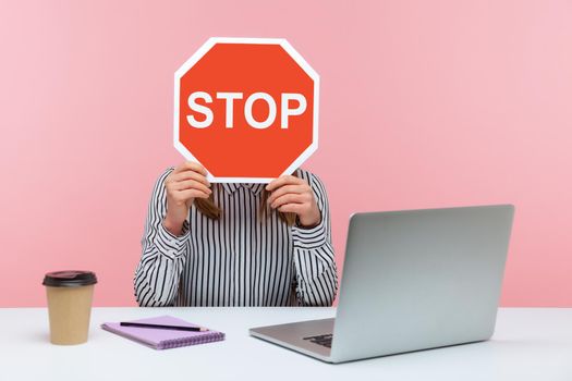 Female office worker in striped shirt sitting at workplace hiding face behind red stop traffic sign avoiding conflicts, afraid of workplace bullying. Indoor studio shot isolated on pink background