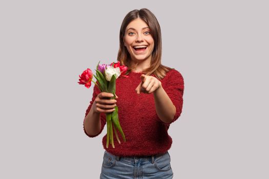 Flowers for you. Portrait of cheerful happy brunette girl smiling, holding tulips bouquet and pointing at camera. Congratulations on spring holidays. indoor studio shot isolated on gray background