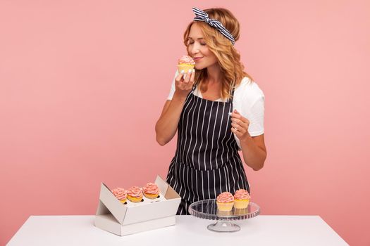 Blonde young adult baker takes out cakes from paper box, preparing for serving to guests, enjoying of smelling tasty dessert with closed eyes. Indoor studio shot isolated on pink background.