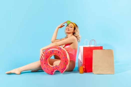 Portrait of joyful glamour girl in swimsuit sitting with rubber ring and shopping bags, looking at camera with toothy smile, enjoying summer sales. indoor studio shot isolated on blue background