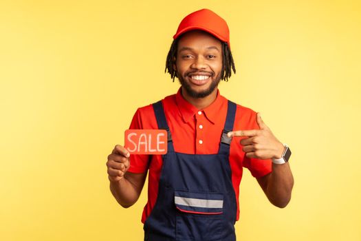 Handsome positive handyman or mechanic wearing red T-shirt and blue uniform, pointing at cad with sale inscription, discounts for service industry. Indoor studio shot isolated on yellow background.