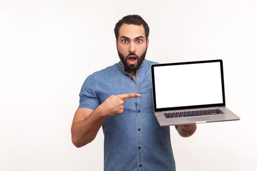 Surprised man pointing finger at empty display of laptop looking at camera with big eyes and opened mouth, shocked with advertisement. Indoor studio shot isolated on white background