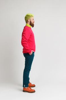 Profile of young serious concentrated bearded man standing with hands in pockets, looking forward, wearing red jumper and pants, has bright hair. Indoor studio shot isolated on gray background.