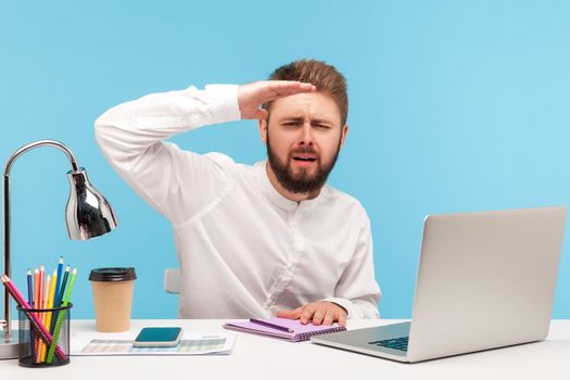 Ambitious bearded man office worker sitting workplace with laptop and looking far away with hand above eyes, searching with promotion, exploring future. Indoor studio shot isolated on blue background