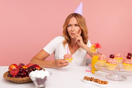 Thoughtful blonde woman wearing party cone thinking about birthday desire, making wish, frowning face and keeps finger in cheek, holds cake with candle. Indoor studio shot isolated on pink background.