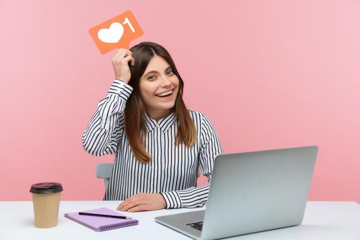 Excited female blogger holding likes counter template above head sitting at workplace with laptop, making posts in social networks. Indoor studio shot isolated on pink background
