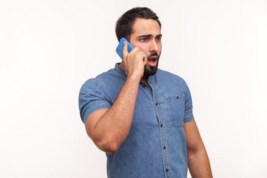 Shocked man with beard in blue shirt talking phone, surprised with news looking big eyes and widely opened mouth, unpleasant call, bad tariffs, roaming. Indoor studio shot isolated on white background
