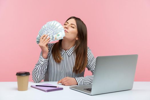 Excited rich business woman enjoying smell of hundred dollar bills holding cash in hand sitting at workplace, crazy about big profit. Indoor studio shot isolated on pink background