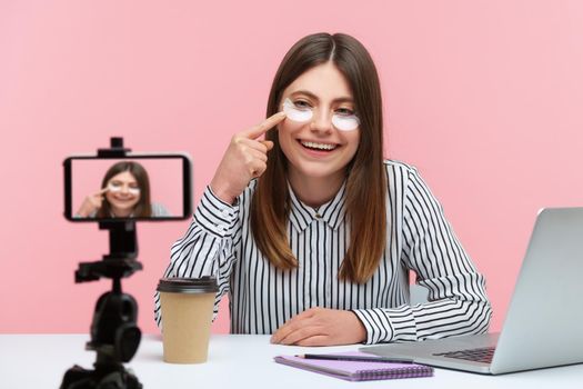 Attractive cheerful woman beauty blogger recording video for followers, testing and recommending eye patches, keeping vlog with makeup tips. Indoor studio shot isolated on pink background