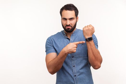 Annoyed punctual man with beard in blue shirt pointing finger at wrist watch mock up display and expressing dissatisfaction with late time, deadline. Indoor studio shot isolated on white background