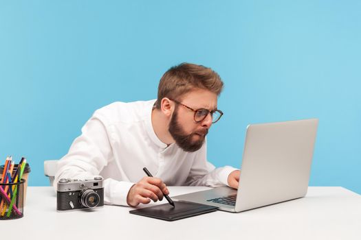 Concentrated bearded man designer drawing on professional tablet looking at laptop display, creating project sitting at workplace. Indoor studio shot isolated on blue background