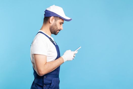 Side view, handyman in overalls typing on mobile phone, using cellphone messenger to accept online order as delivery, repair and maintenance services. indoor studio shot isolated on blue background