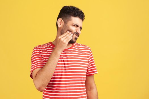 Unhappy bearded man in red striped t-shirt feeling toothache, touching sore cheek, suffering from cavities, cracked teeth, gum recession. Indoor studio shot isolated on yellow background