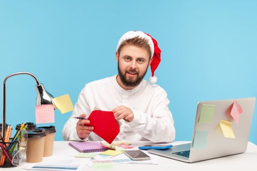 Handsome bearded man office worker in santa claus hat sitting at workplace and signing postcards, congratulating with winter holidays, making surprise. Indoor studio shot isolated on blue background