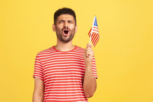 Devoted patriotic man in striped t-shirt holding in hand flag of united states of america, singing hymn celebrating independence day. Indoor studio shot isolated on yellow background