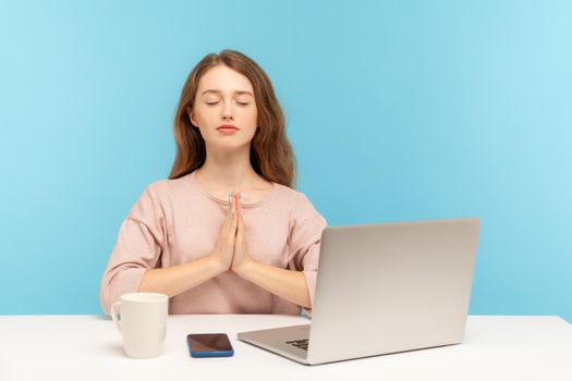 Break at work, yoga practice. Calm woman employee sitting at workplace with laptop and raising hands in namaste gesture, meditating at home office. indoor studio shot isolated on blue background