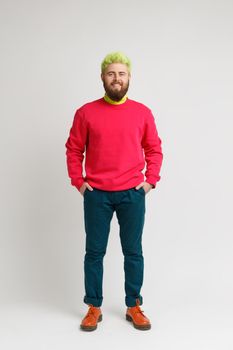 Full length photo of happy smiling bearded man with bright appearance, wearing red sweater, pants and shoes, keeps hands in pockets. Indoor studio shot isolated on gray background.