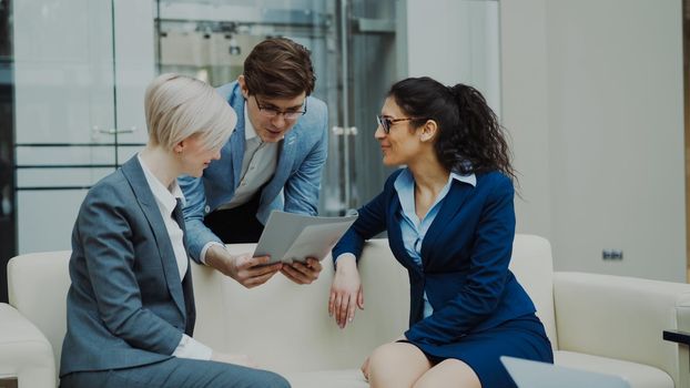 Cheerful businessman talking and discussing about financial report with business female colleagues sitting on sofa in meeting room indoors