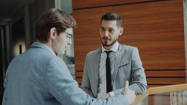 Two businessman colleagues standing and chatting standing near railing in hall of modern business center indoors