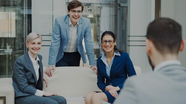 Team of cheerful business colleagues talking and discussing each other while having break sitting on sofa in modern office hall indoors