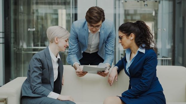 Cheerful businessman talking and discussing about financial report with business female colleagues sitting on sofa in meeting room indoors