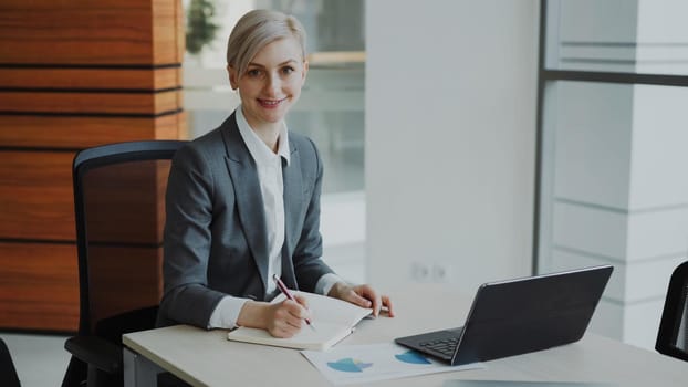 Portrait of Attractive blonde businesswoman sitting at table writing in notebook smiling into camera in modern office indoors