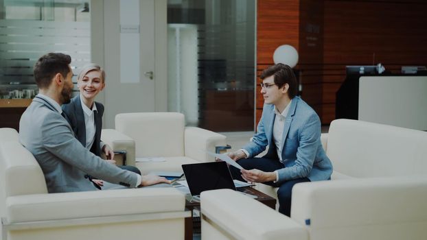 Cheerful businessman discussing financial reports with business partners sitting on sofa in modern office hall indoors