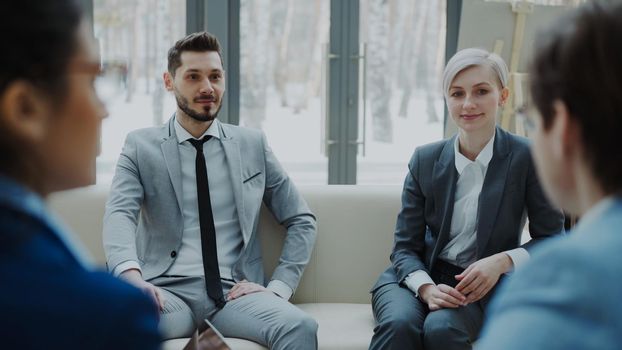 Cheerful businessman and businesswoman talking and duscussing future contract with business partners sitting on couch in meeting room indoors
