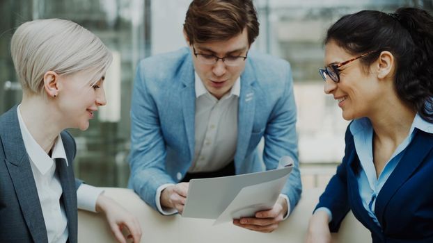 Close-up of cheerful businessman talking and discussing about financial report with business female colleagues sitting on sofa in meeting room indoors