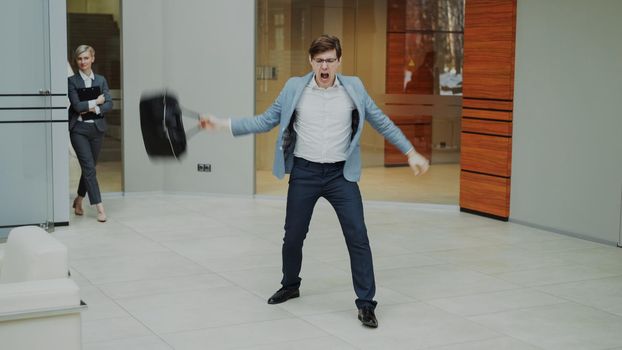 Crazy businessman dancing with briefcase in modern lobby indoors while his colleagues walking and watching him surprised