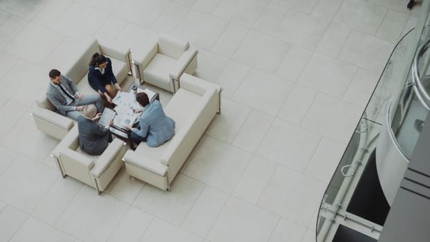 Top view of group of business people colleagues discussing financial charts sitting on couchs in lobby at modern business center indoors