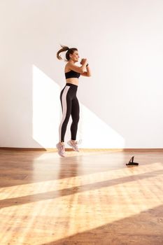 Female jumping, doing cardio exercises after fitness blogger, watching tutorial videos on tablet, wearing black sports top and tights. Full length studio shot illuminated by sunlight from window.