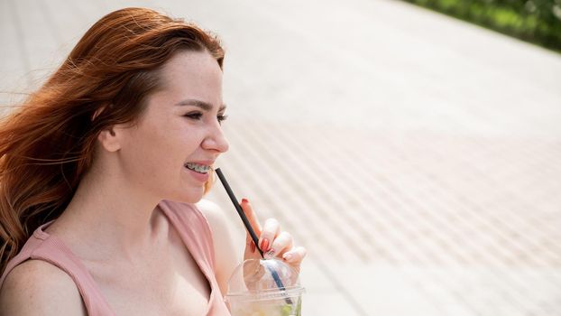 Young beautiful red-haired woman with braces drinks cooling lemonade outdoors in summer. Portrait of a smiling girl with freckles