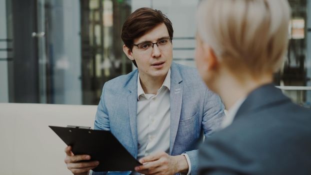 HR manager with clipbooard interviewing young female candidate for hiring on vacant position sitting on sofa in modern office indoors