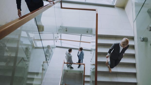 Top view of two businessmen meet at staircase in modern office center indoors and talking while female colleagues walking stairs