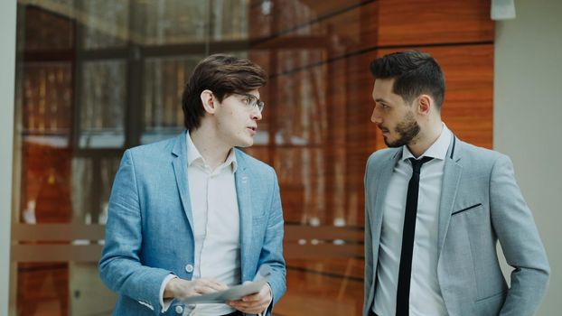 Two businessmen colleagues walking and discussing documents in modern office hall indoors