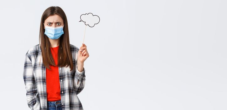 Coronavirus outbreak, leisure on quarantine, social distancing and emotions concept. Troubled young woman in medical mask frowing upset or disappointed, hold comment cloud stick near head.