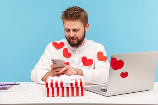 Smiling bearded businessman in white shirt in red heart shaped stickers sitting at desk with laptop and striped giftbox typing on smartphone. Indoor studio shot isolated on blue background