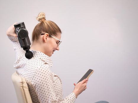 A young woman sits on a chair, uses a smartphone and makes herself massage with an electro massage gun