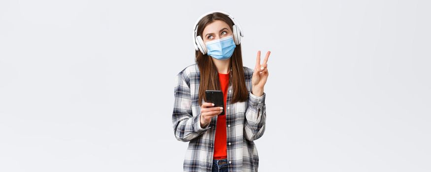 Social distancing, leisure and lifestyle on covid-19 outbreak, coronavirus concept. Happy cheerful silly girl in medical mask and headphones, show peace sign smiling, holding mobile phone.