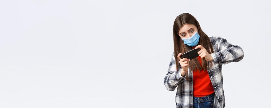 Different emotions, covid-19 pandemic, coronavirus self-quarantine and social distancing concept. Focused and entertained woman in medical mask playing mobile game, tilting body, difficult level.